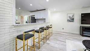 A modern kitchen with white cabinets, a marble countertop, and a built-in oven. There are four bar stools with black cushions and gold frames. A flat-screen TV is mounted on the wall, and a framed picture hangs above it. - enjoy ranch life.

 (Allen)