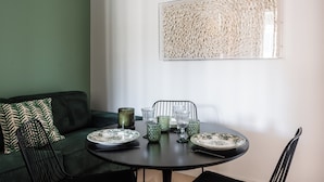 Un comedor con una mesa redonda dispuesta para cuatro personas, rodeada de sillas tapizadas de verde. Un cojín verde estampado se encuentra en el sofá a la izquierda.