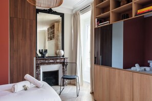 Un dormitorio con cama, una silla, chimenea, armario y un espejo grande.