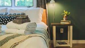 Un lit bien fait, avec des serviettes pliées, une table de chevet avec une lampe et une petite plante, et une fenêtre avec des rideaux.