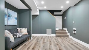 Un salon moderne avec un mur gris foncé, un canapé gris agrémenté de coussins à motifs, un plancher en bois et un escalier menant à un autre niveau.