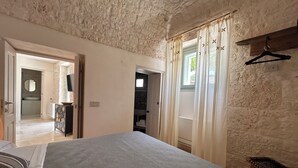 A bedroom with a large bed, a wooden bench, a hanging clothes rack, and a view of a bathroom and a kitchen through an open door. - Charming suites among trulli in the heart of Alberobello (Alberobello)