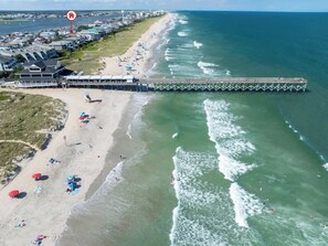 An aerial view of a beach with a pier extending into the ocean, beach umbrellas, and a residential area with houses.