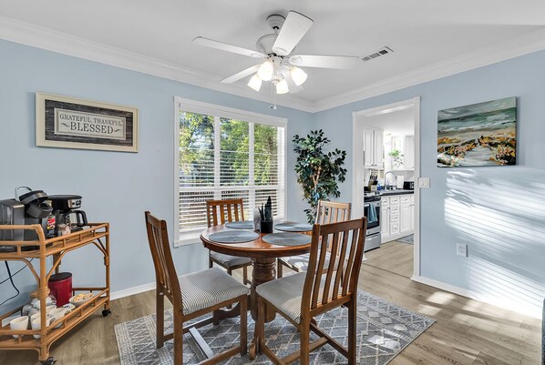 A dining area with a round table, four chairs, a ceiling fan, and a kitchen in the background.