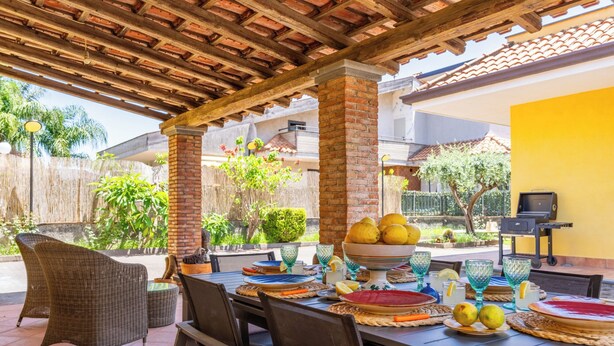 An outdoor patio with a wooden pergola, wicker chairs, a table set with plates, glasses, and lemons. - Villa Cesare (Mascali)