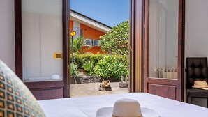 A hotel room with a bed, a hat on the bed, and a view of the outdoor area with plants and a building in the background. - Villa Cesare (Mascali)