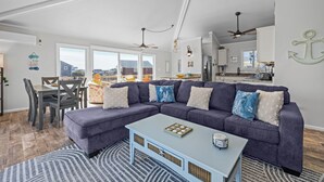 En moderne stue med en modulsofa, spisebord og stole. Der er et køkkenområde med skabe og hårde hvidevarer i baggrunden.