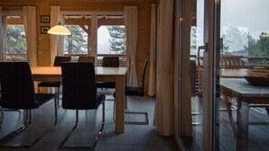 Een houten interieur met een eethoek, grote ramen en uitzicht op bomen en bergen.