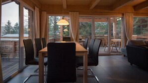 Een houten interieur met een eettafel en stoelen, grote ramen en uitzicht op bomen.