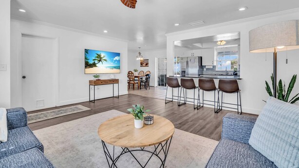 Un salon spacieux avec une table basse en bois, un canapé et un ventilateur de plafond. On aperçoit une cuisine avec des tabourets de bar à l’arrière-plan.