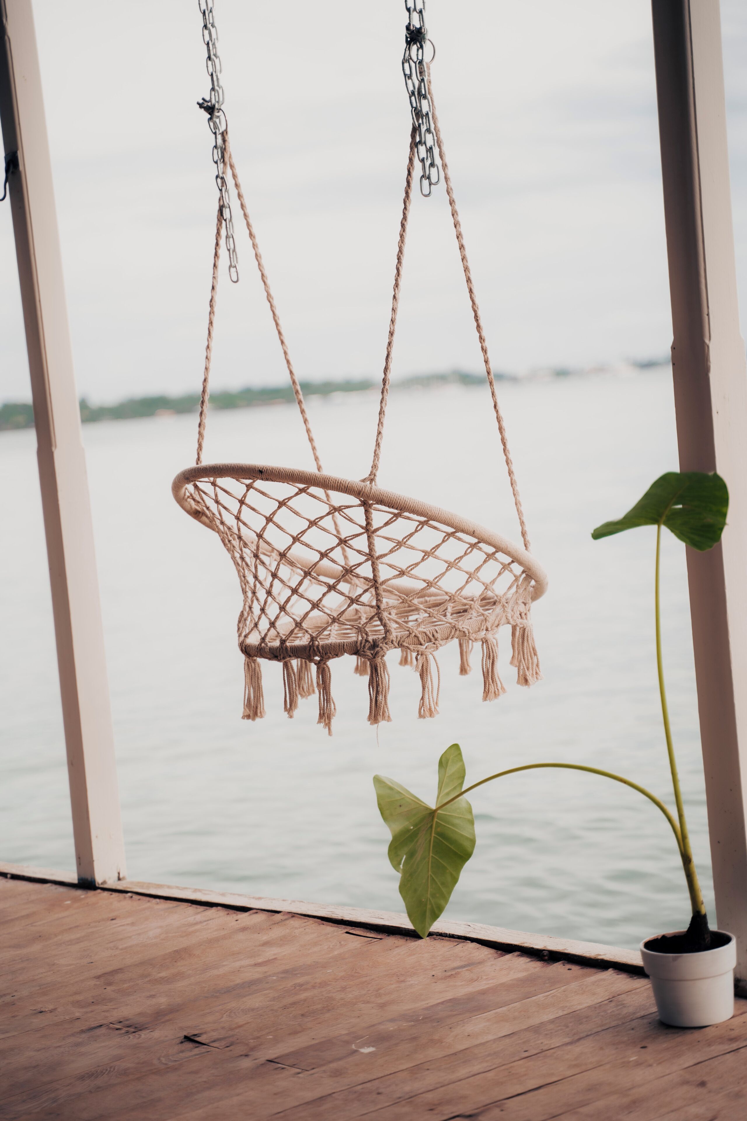 Un fauteuil suspendu en macramé, maintenu par des chaînes, une plante en pot et une terrasse en bois offrant une vue sur l’eau.