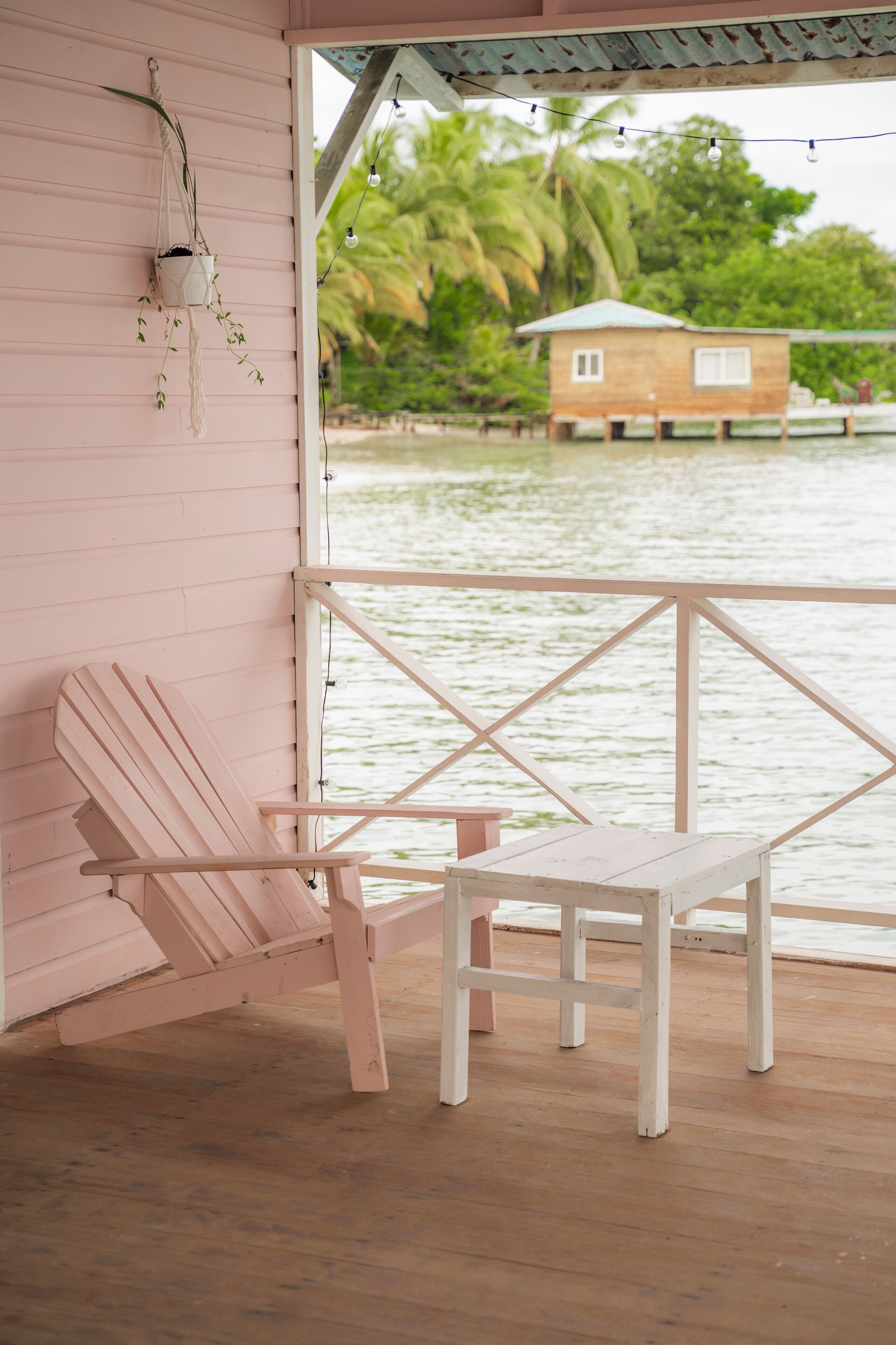 Une terrasse en bois avec une chaise rose et une petite table, donnant sur une étendue d’eau, avec une maison en bois en arrière-plan.