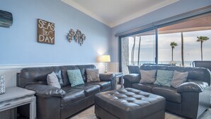 A living room with two dark leather sofas, a small white table, and a large window overlooking a beach.