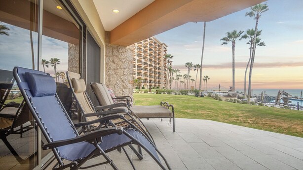 A hotel balcony with lounge chairs, a stone wall, and a view of a high-rise building and palm trees.
