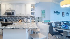 A modern kitchen with white cabinetry, a granite countertop, and a built-in microwave. There is a dining area with a table and chairs, and decorative wall art.