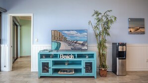 A living room with a teal console table labeled "Beach," a large framed picture of a beach resort, a bamboo plant, and a refrigerator.