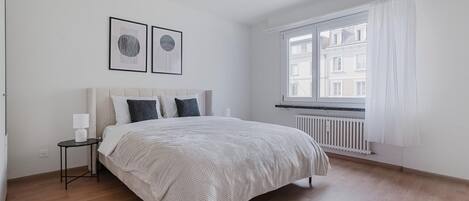A bedroom with a bed, a nightstand, a window with curtains, and a radiator.