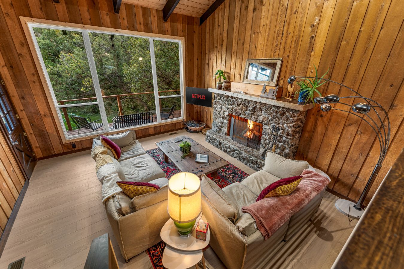 A cozy cabin interior with a stone fireplace, a sofa set, and a large window overlooking a forest.