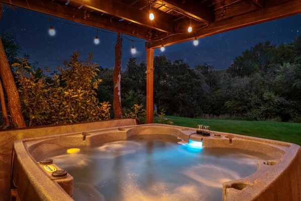 An outdoor hot tub with a wooden pergola overhead, surrounded by trees and a night sky.