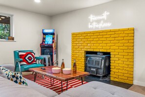 A living room with a yellow brick wall, a neon sign, a retro arcade machine, a sofa, a coffee table, and a fireplace. - Hot Tub|Riverfront|15 min to Hot Springs & Skiing (Glenwood Springs)