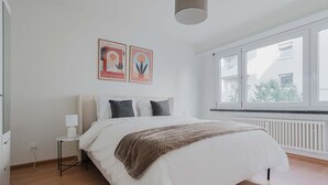 A bedroom with a bed, a bedside table with a lamp, a radiator, and two framed artworks on the wall.