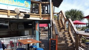 Un espace extérieur aménagé pour les repas, avec des tables en bois et des tabourets bleus, une enseigne indiquant « Accueil », et un escalier menant à un niveau supérieur.