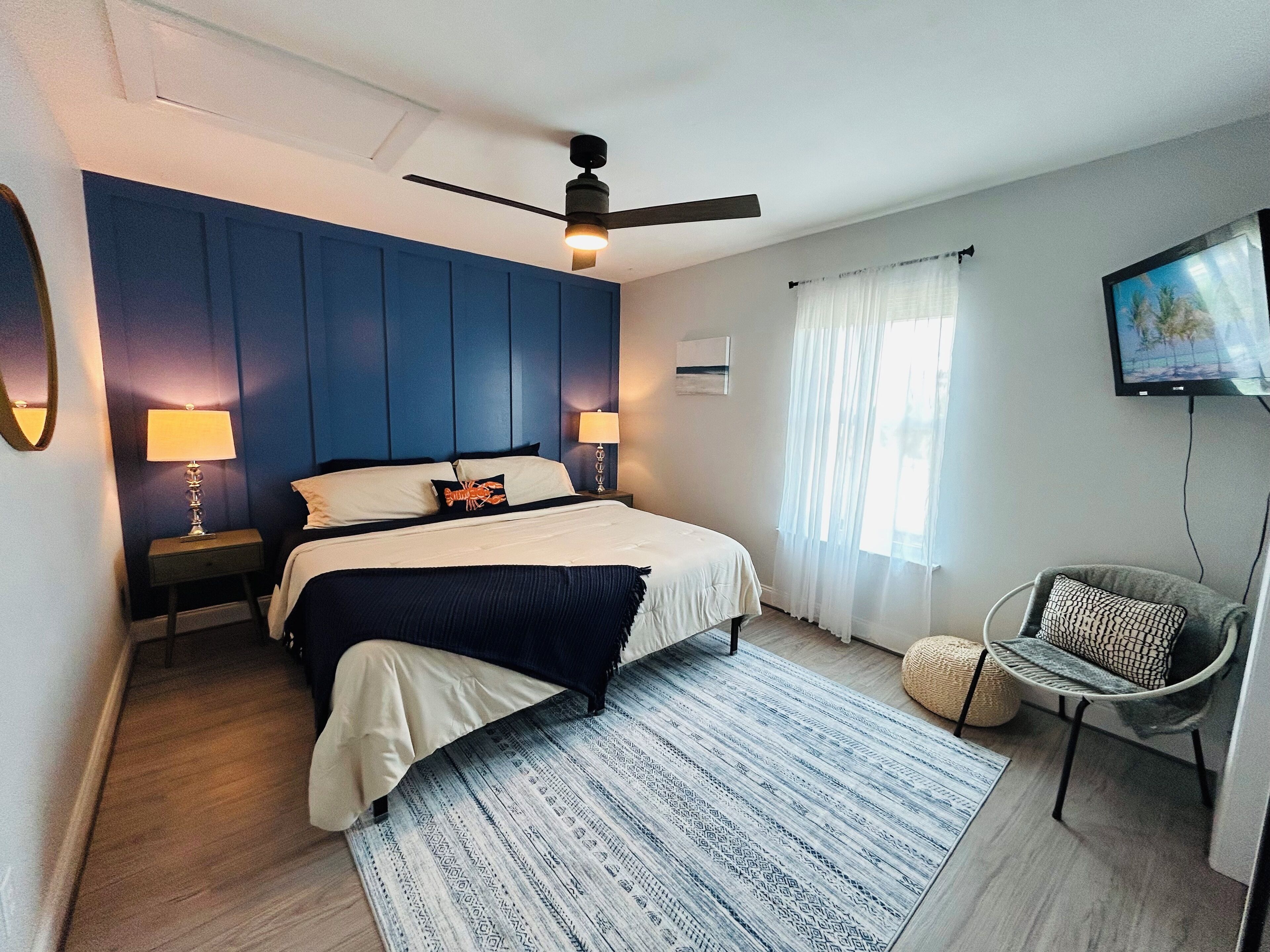 Un dormitorio moderno con una cama, dos mesitas de noche con lámparas, un ventilador de techo, un televisor en la pared y una silla con cojín.