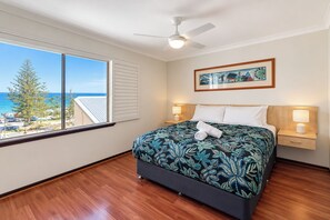 Een hotelkamer met een bed, twee bedlampen, een plafondventilator, een raam met uitzicht op de oceaan en een ingelijst kunstwerk aan de muur.