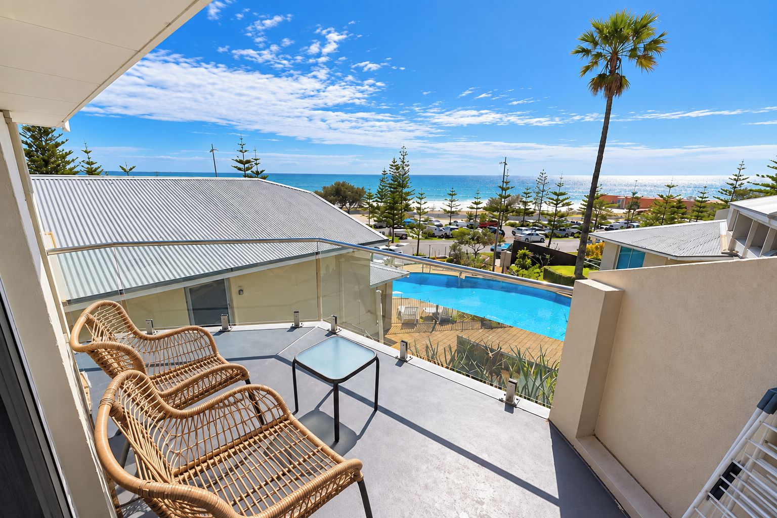 A balcony with wicker chairs and a glass table overlooking a swimming pool and a coastal view.