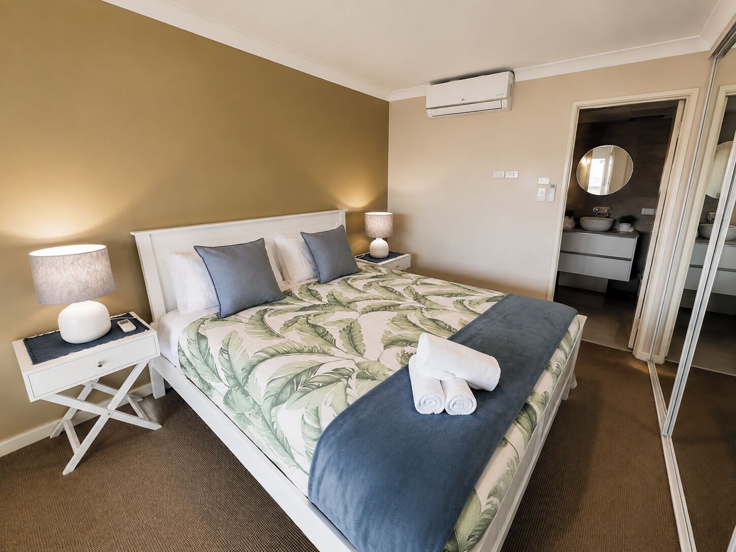 A hotel room with a bed, bedside tables, lamps, an air conditioning unit, a bathroom with a mirror, and a towel on the bed.