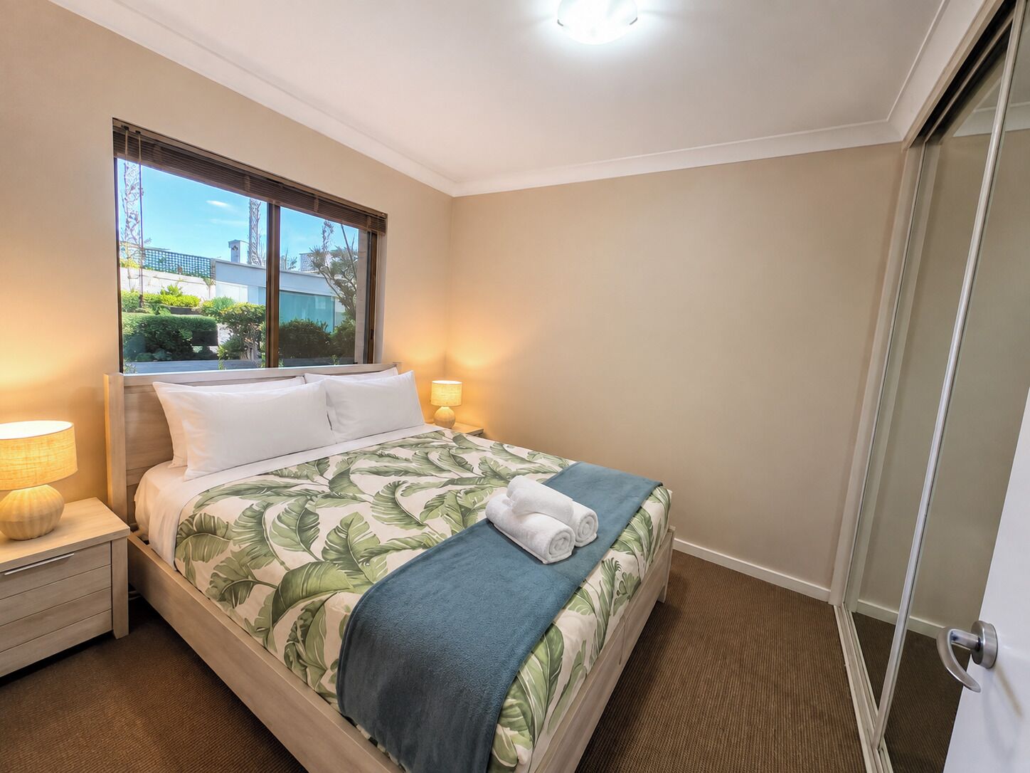 Una habitación de hotel con una cama, una mesita de noche, una lámpara, una ventana con persianas y una puerta.