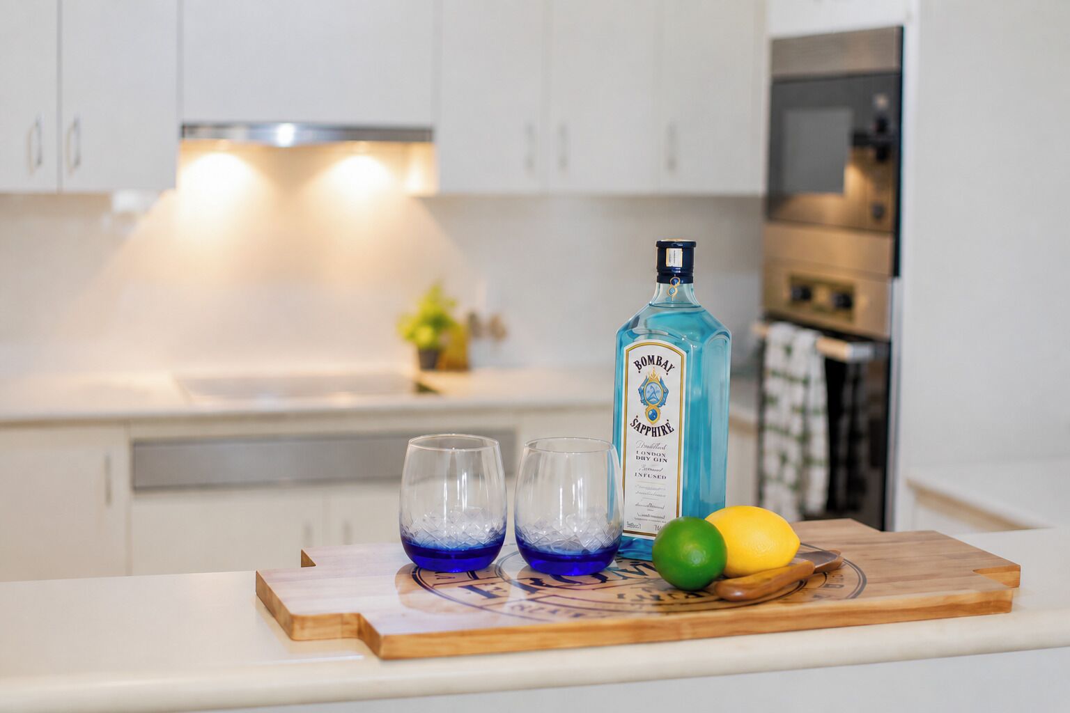 A kitchen counter with a bottle of Bombay Sapphire gin, two glasses with ice, and a wooden cutting board with lemons and a spoon.