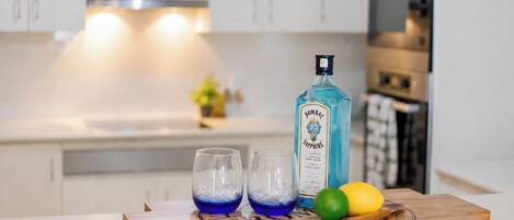 A kitchen counter with a bottle of Bombay Sapphire gin, two glasses with ice, and a wooden cutting board with lemons and a spoon.