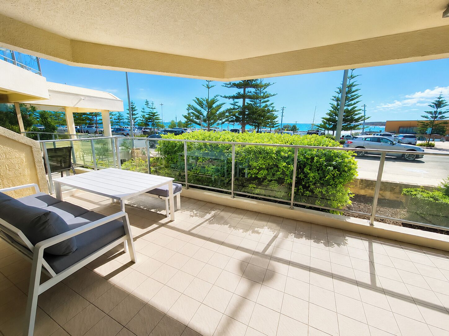 A balcony with a white table and two chairs, a glass railing, and a view of a parking lot and trees.