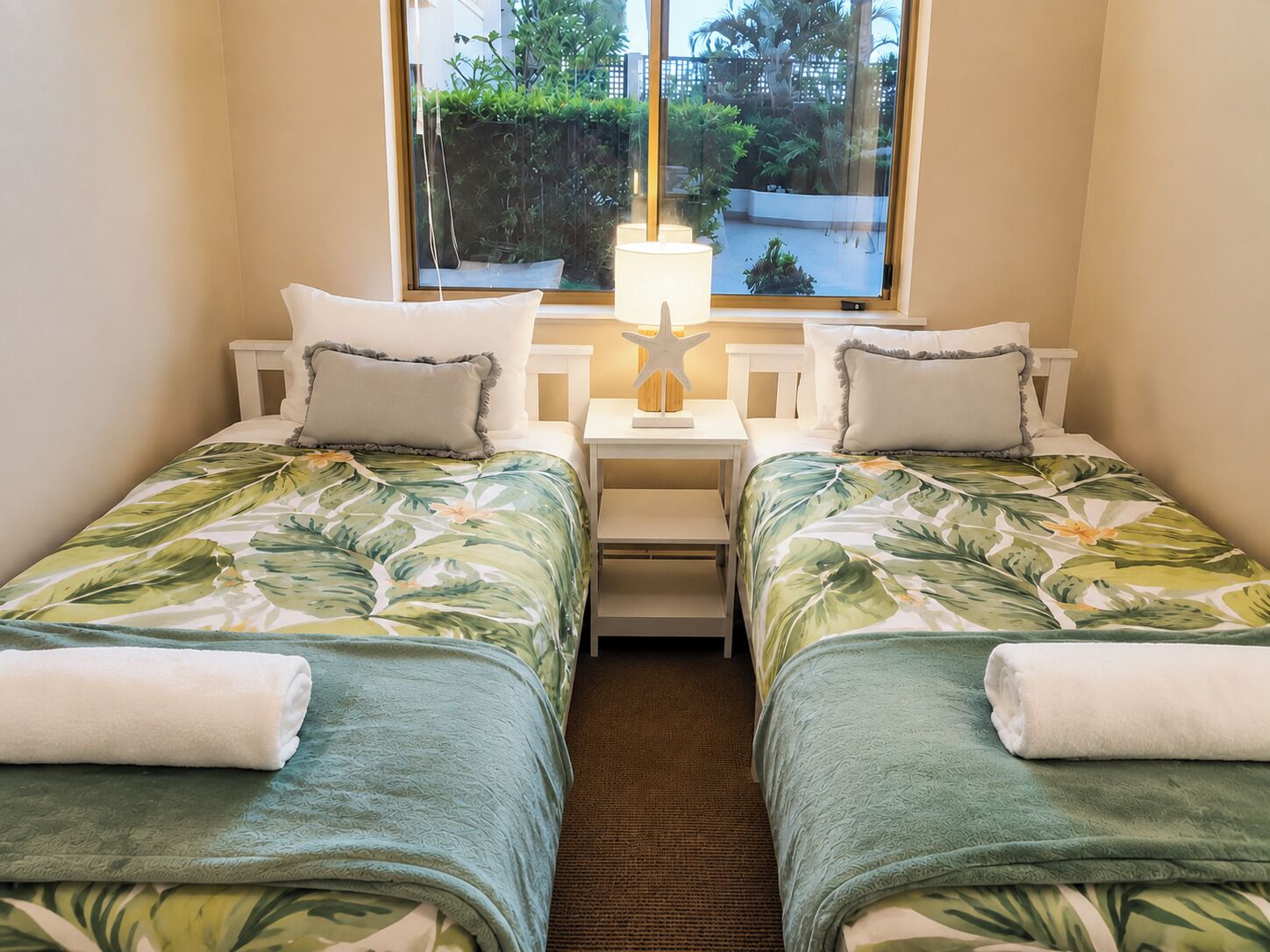 Ein Hotelzimmer mit zwei Einzelbetten, einem Nachttisch mit Lampe und einem Fenster mit Blick auf Grünflächen.