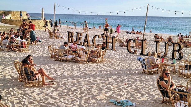 A beach club with wicker chairs and tables, a stone wall, and the ocean in the background. - Sunset Shores Scarborough (Scarborough)