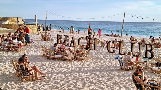 Un club de plage avec des chaises et des tables en osier, un mur de pierre et l’océan en toile de fond.