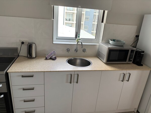 A compact kitchen with a stainless steel sink, a kettle, a microwave, and a window with a view of buildings.