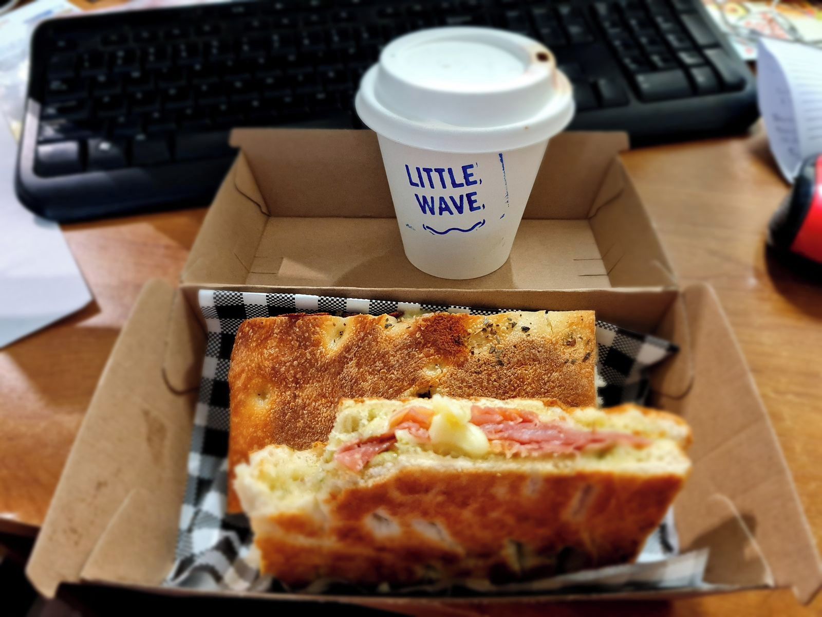 A sandwich in a box with a coffee cup labeled 'Little Wave' on a desk.