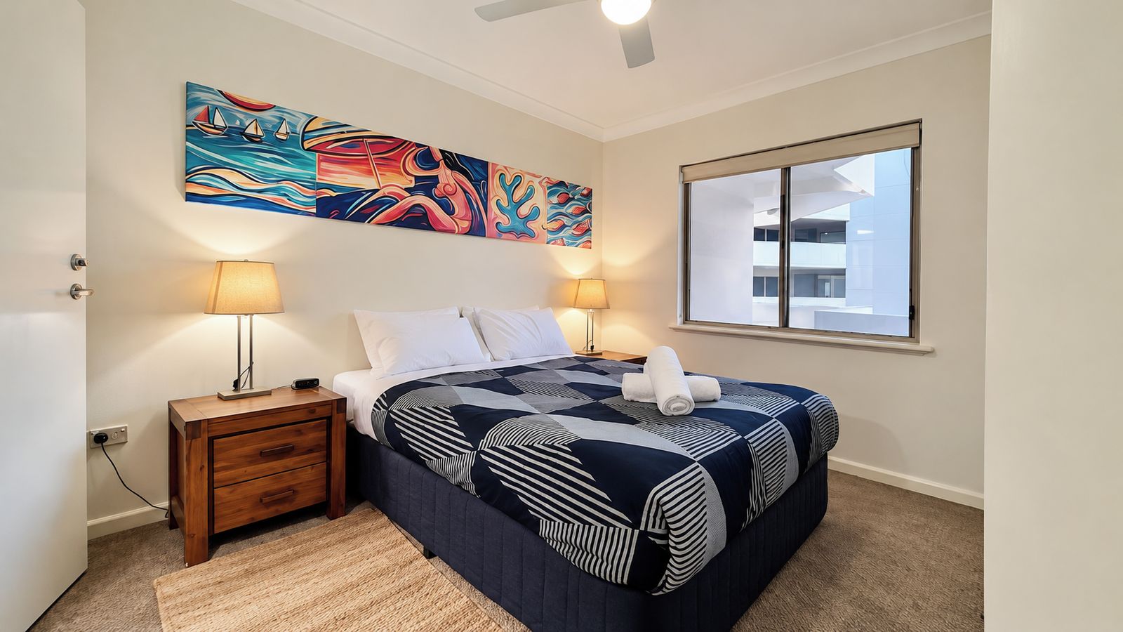 Une chambre à coucher avec un lit, deux lampes de chevet, une table de nuit, une grande fenêtre et un tableau abstrait aux couleurs vives accroché au mur.