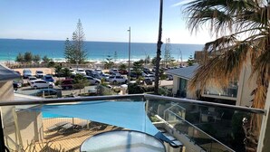 A balcony with a glass table and chairs overlooking a pool and a parking area with cars and a beach in the background. - Scarborough Sunset on Esplanade (Scarborough)