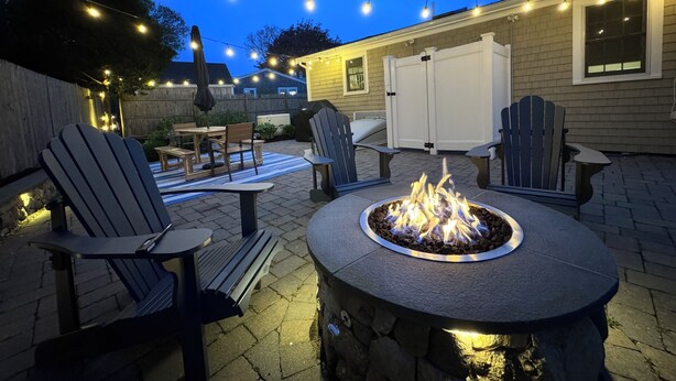 Un patio al aire libre con sillas tipo Adirondack, una fogata y luces colgantes.