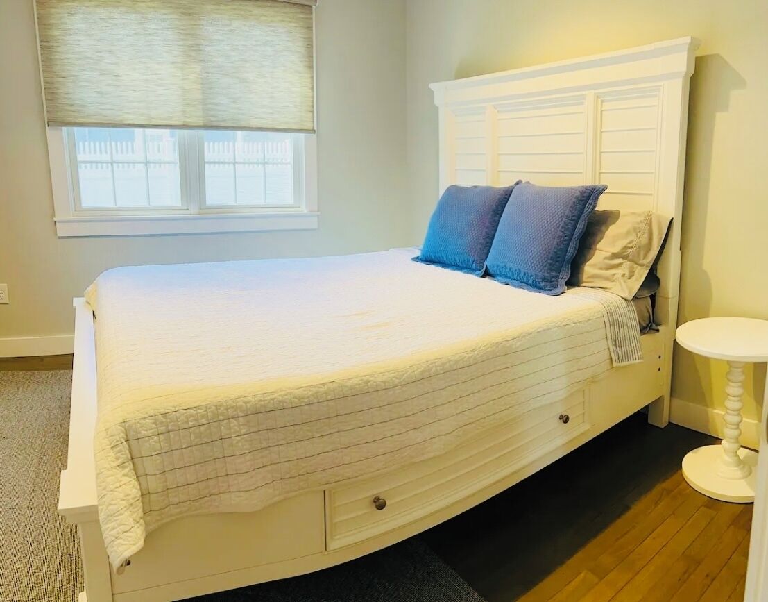 A neatly made bed with a white headboard and two pillows, a small bedside table, and a window with blinds.