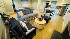 Una sala de estar moderna con un sofá gris, sillones negros y un otoman redondos. Se ve una cocina con armarios blancos y electrodomésticos de acero inoxidable al fondo.