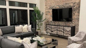 Ein modernes Wohnzimmer mit einem schwarzen Ledersofa, grauen Sesseln, einem Glastisch und einer Ziegelwand mit einem Flachbildfernseher.
