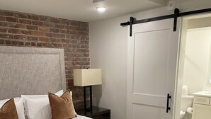 Ein Schlafzimmer mit einem großen Bett, einer Wand aus Ziegelsteinen, einem Deckenventilator, einem Nachttisch mit Lampe und einer Schiebetür zu einem Badezimmer.