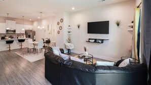 Een moderne woonkamer met een zwarte leren bank, een eethoek met een witte tafel en stoelen, een keuken met witte kasten en een bar, en een wandgemonteerde flatscreen-televisie.