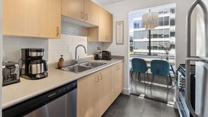 Una cocina moderna con armarios de madera, electrodomésticos de acero inoxidable y lavavajillas. Se ve una ventana con vista a un edificio y una lámpara de araña sobre el fregadero.