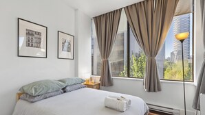 Una habitación de hotel moderna con una cama grande, dos cuadros en la pared, una mesita de noche y vistas a un paisaje urbano por la ventana.