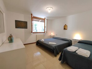 Une chambre avec deux lits, une fenêtre, un radiateur, une table de chevet avec une lampe et un petit objet décoratif sur une commode.